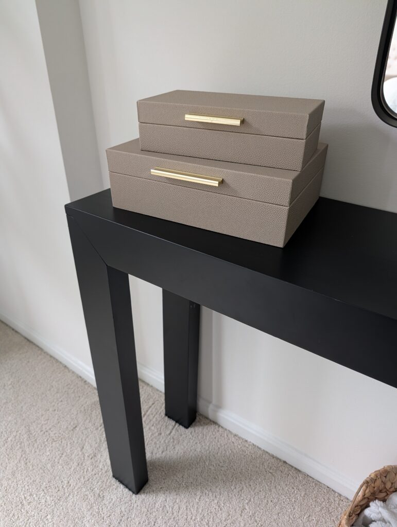 In this photo you see the edge of a simple modern entry table in black. Angled on the corner is a stack of two boxes with metal handles.