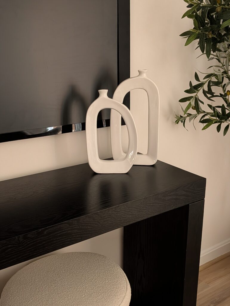 This pictures shows an entryway table in simple ebony wood. Two modern white vases sit on the table and a storage ottoman sits below. To the right is a faux plant bringing in some greenery to the corner.