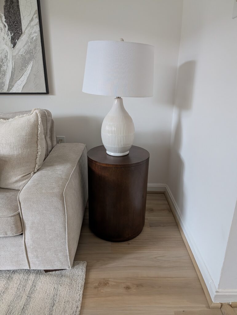 This image shows a minimalist cylindrical end table in a dark warm wood tone sitting next to a light beige sofa. On the end-table is a single white ceramic table lamp with traditional white shade.