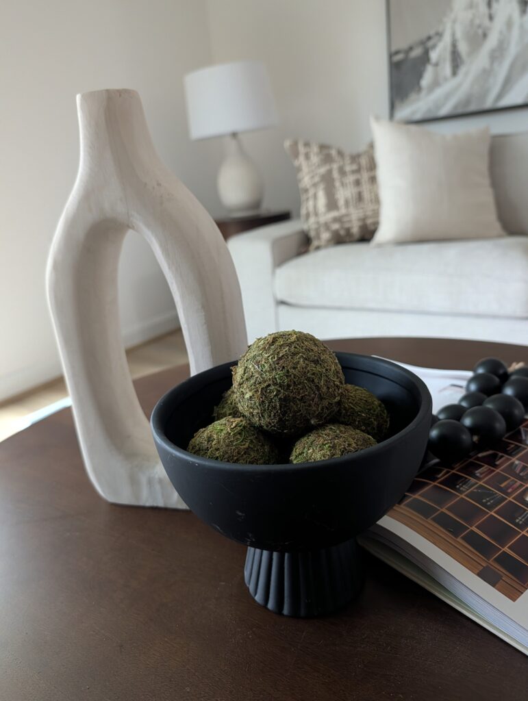 This image is a close-up of a coffee table vignette. On the table is an oval boho vase in white ceramic, a black serving bowl filled with decorative moss balls, an open book with some oversized wooden beads on a string draped over the center.