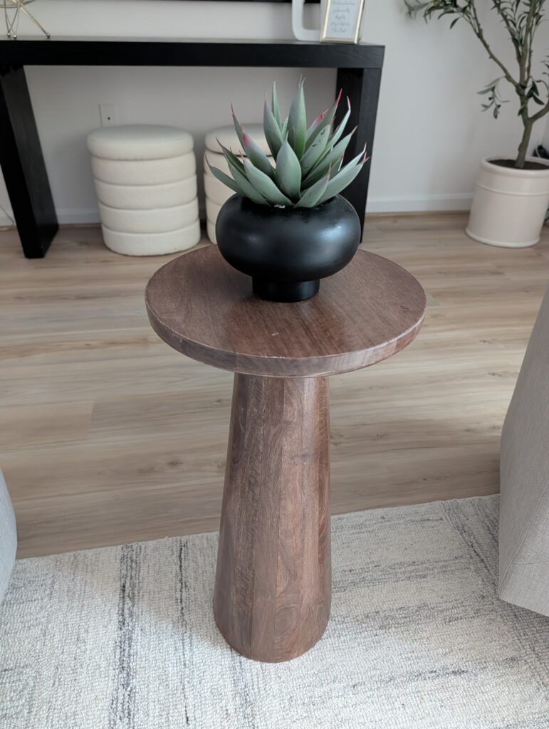 In this photo you see a single pedestal table in warm wood used as a side table for an accent chair. On the table is a single potted plant in a black pot.