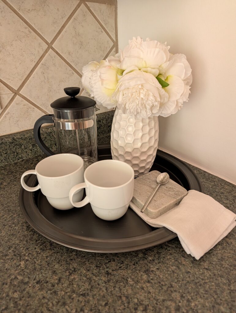 Image shows a staged vignette for a kitchen counter with a french press, a white ceramic vase of white flowers, two coffee mugs, a kitchen towel, a couple of stone coasters arranged on a round tray.
