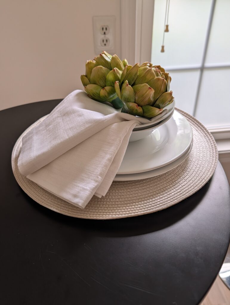 This staging photo shows a woven charger, two stacked white plates with two stacked white bowls. In the top bowl, two kitchen flour sack napkins are arranged draped over the side and held in place by two faux artichokes.