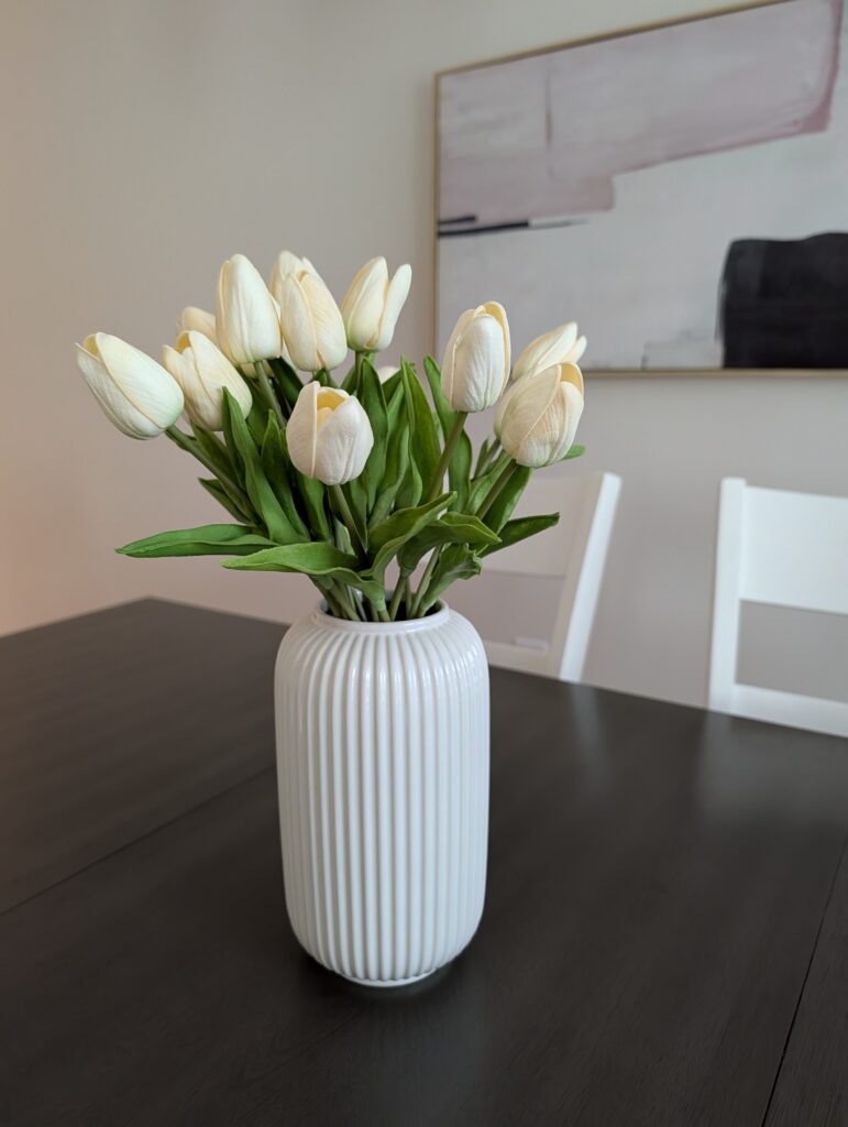 This staging photo shows a dining table with a simple textured white vase filled with white tulips. In the background is a neutral piece of modern geometric art on the wall.