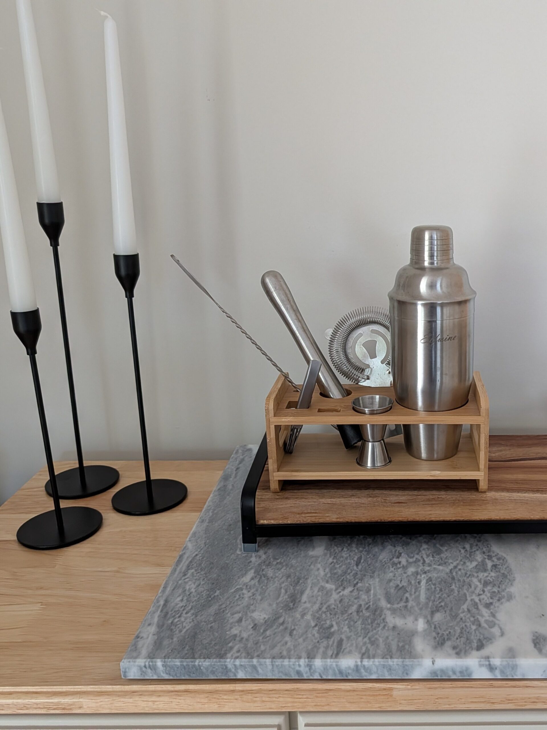 This image is from a console table where a stone cutting board sits layered with a wooden tray, on which sits a cocktail shaker kit. To the left are 3 simple metal candlesticks of varying height.