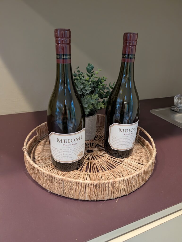This staging photo shows a woven basket tray with a small faux succulent in a white ceramic pot and two wine bottles in a kitchen vignette.