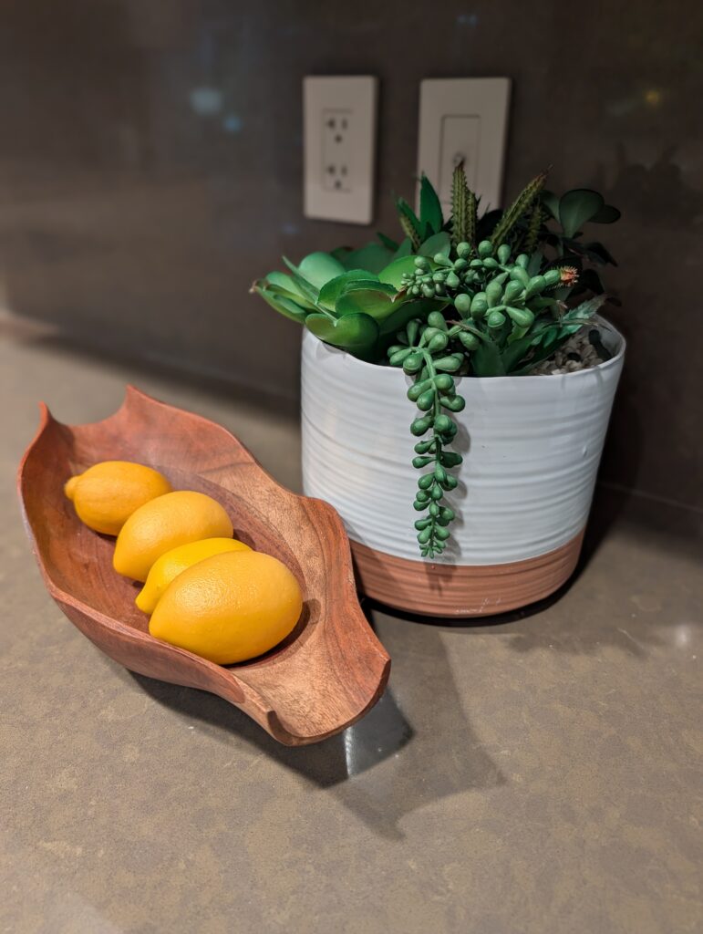 This photo shows a raw edge wooden serving bowl filled with yellow lemons and a white and tan ceramic pot filled with a faux succulent.